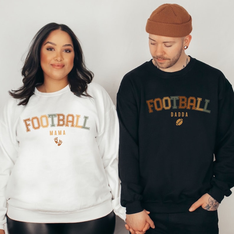 May include: A white sweatshirt with the text "FOOTBALL MAMA" in retro-style lettering, with small footprints below. A black sweatshirt with "FOOTBALL DADDA" in similar lettering and a football graphic. Both are worn by a couple.