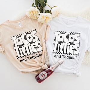 May include: Two white t-shirts with the text "Tacos Titties and Tequila!" printed in black. The shirts are laid out on a white surface with a bottle of sparkling wine.