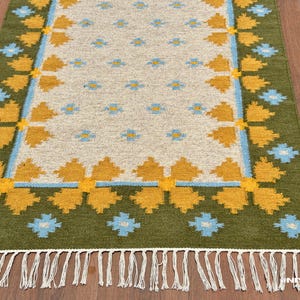 May include: Rectangular rug with a cream-colored center and a green border. The rug features a repeating pattern of yellow and blue floral designs. White fringe trims the bottom edge. The rug is placed on a wooden floor.