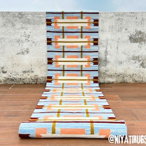 May include: A long rectangular runner rug featuring a light blue background with a repeating geometric pattern of peach-colored rectangles, white horizontal bars, and thin vertical stripes in mustard yellow and dark maroon brown colors.