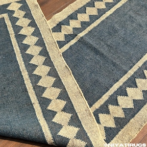 Könnte beinhalten: Ein rechteckiger blauer und beiger Teppich mit einem geometrischen Rautenmuster. Der Teppich hat eine gewebte Textur und einen Fransenrand. Das Design zeigt einen Rand und Rautenformen in Beige auf blauem Hintergrund.