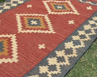 May include: A rectangular red rug with a geometric pattern. The rug has a border of dark gray and beige floral designs. The central design features diamond shapes with blue, yellow, and beige accents. The rug is on a green grassy surface.