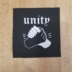 Könnte beinhalten: Schwarzer quadratischer Aufnäher mit dem Wort „unity“ in weißer Schrift über einer weißen Grafik von Händedruck. Der Aufnäher, wahrscheinlich aus Stoff, liegt auf einer Holzoberfläche.