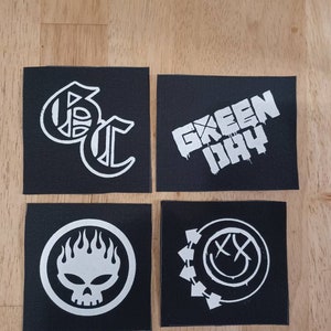 Può includere: Quattro toppe di tessuto nero con loghi di band bianchi. Le toppe presentano i nomi delle band "Green Day", "Offspring", "Bad Religion" e "blink-182".