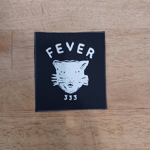 Puede incluir: Parche de tela negro con texto blanco que dice "FEVER" y un gato de dibujos animados blanco con el número "333" debajo.