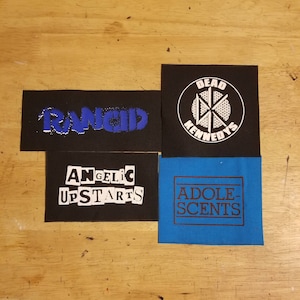 以下が含まれることがあります： バンドのロゴが入った4つの布パッチ。左上のパッチは黒で、青色の「RANCID」という文字が入っています。右上のパッチは黒で、白い「DEAD KENNEDYS」のロゴが入っています。左下のパッチは黒で、白い「ANGELIC UPSTARTS」という文字が入っています。右下のパッチは青色で、茶色の「ADOLESCENTS」という文字が入っています。