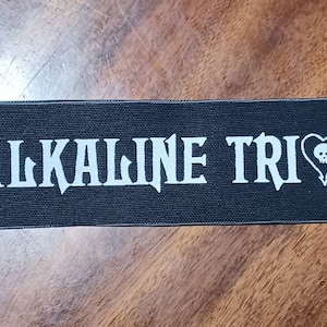 Könnte beinhalten: Schwarzer Stoff-Patch mit weißem Text "ALKALINE TRIO" und einem weißen Herz mit einem Totenkopf darin.