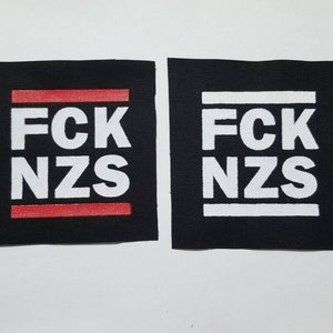 May include: Two black square patches with white text reading "FCK NZS". One patch has a red horizontal line above the text and another red line below the text. The other patch has white lines above and below the text.