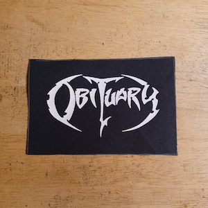 Könnte beinhalten: Schwarzer rechteckiger Aufnäher mit dem Wort "Obituary" in weißer, stilisierter, gotischer Schrift. Der Aufnäher liegt auf einer Holzoberfläche.