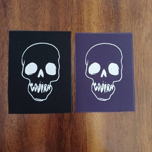 May include: Two fabric patches with a white skull and crossbones design. The skull is outlined in white and has two eye sockets and a nose. The text "GOJIRA" is written below the skull. One patch is black and the other is purple.