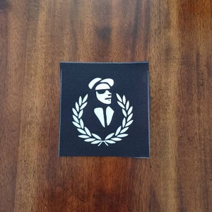 Op de afbeelding: Zwart-witte patch met een wit silhouet van een persoon met een hoed en zonnebril, omringd door een lauwerkrans.