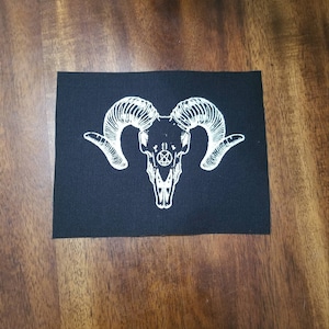 May include: Black rectangular patch featuring a white ram skull design with large curved horns and a pentagram symbol. The patch is made of a woven fabric and is likely intended for application on clothing or accessories.
