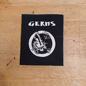 Puede incluir: Parche bordado en blanco y negro con la palabra "GERMS" en letras blancas y un círculo que contiene una calavera y huesos cruzados.