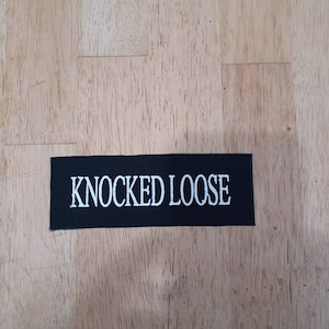 Puede incluir: Un parche rectangular negro con el texto blanco "KNOCKED LOOSE". El parche está hecho de tela tejida y está diseñado para ser cosido en ropa o accesorios. El texto está en negrita y sin serifa.