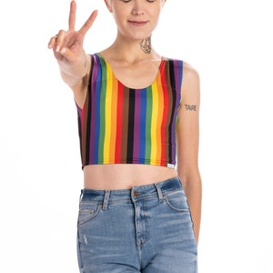 May include: A person wearing a rainbow striped crop top with a black and brown stripe. The person is wearing blue jeans and has short blonde hair. The person is making a peace sign with their hand. The word "TAIRE" is tattooed on their arm.