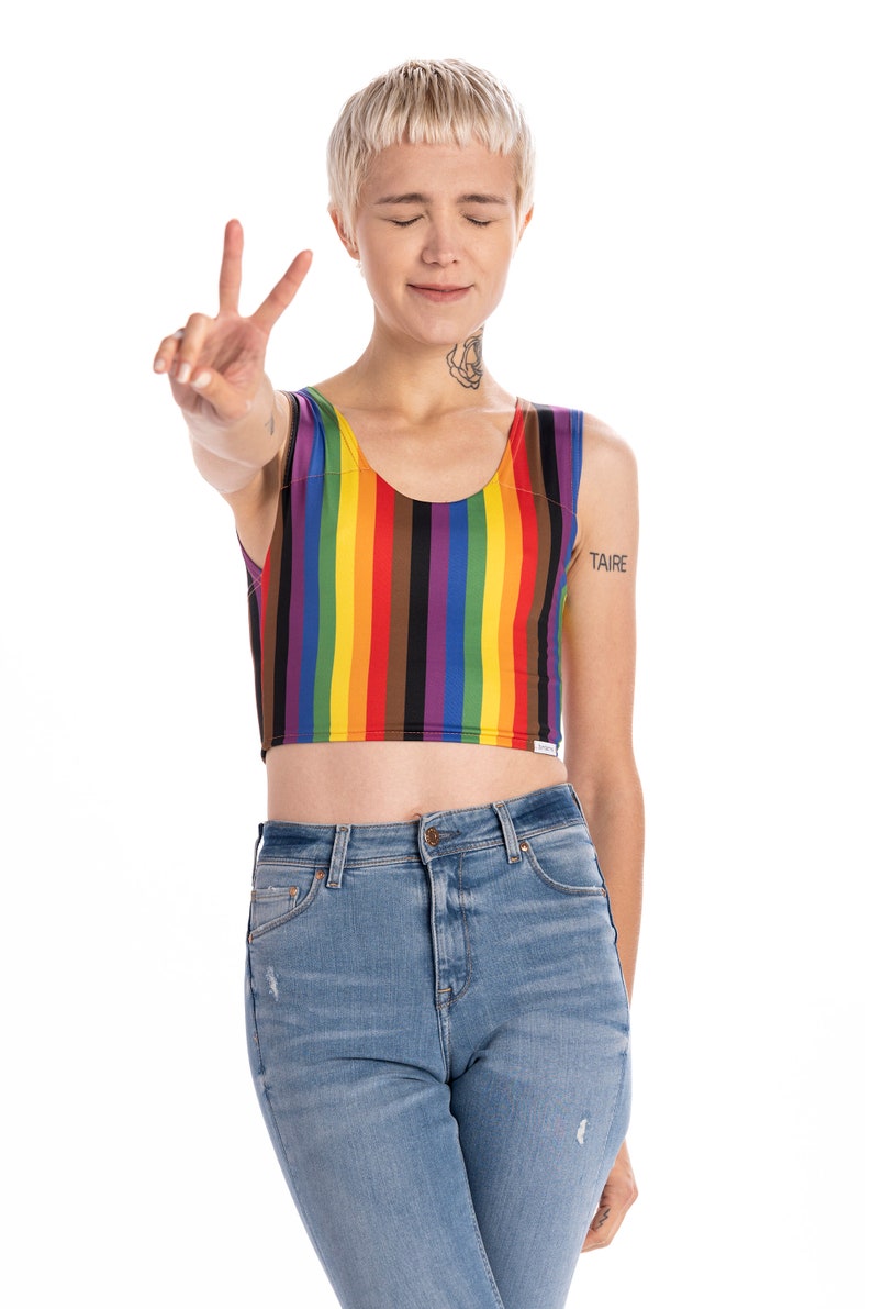 May include: A person wearing a rainbow striped crop top with a black and brown stripe. The person is wearing blue jeans and has short blonde hair. The person is making a peace sign with their hand. The word "TAIRE" is tattooed on their arm.