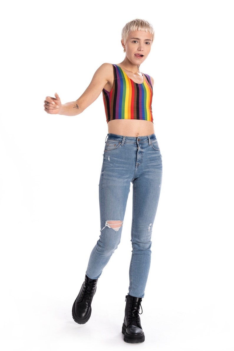 May include: A person wearing a rainbow striped crop top and blue jeans with distressed knees. They are wearing black combat boots.