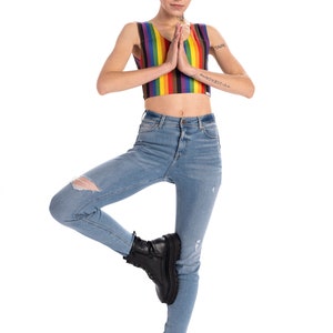 May include: A person wearing a rainbow striped crop top and blue jeans with rips. They are standing on one leg with their other leg bent at the knee and their foot resting on their thigh. They are wearing black combat boots and have tattoos on their arms. The text on their arm reads "TAIPEI" and "PLUS337-A".