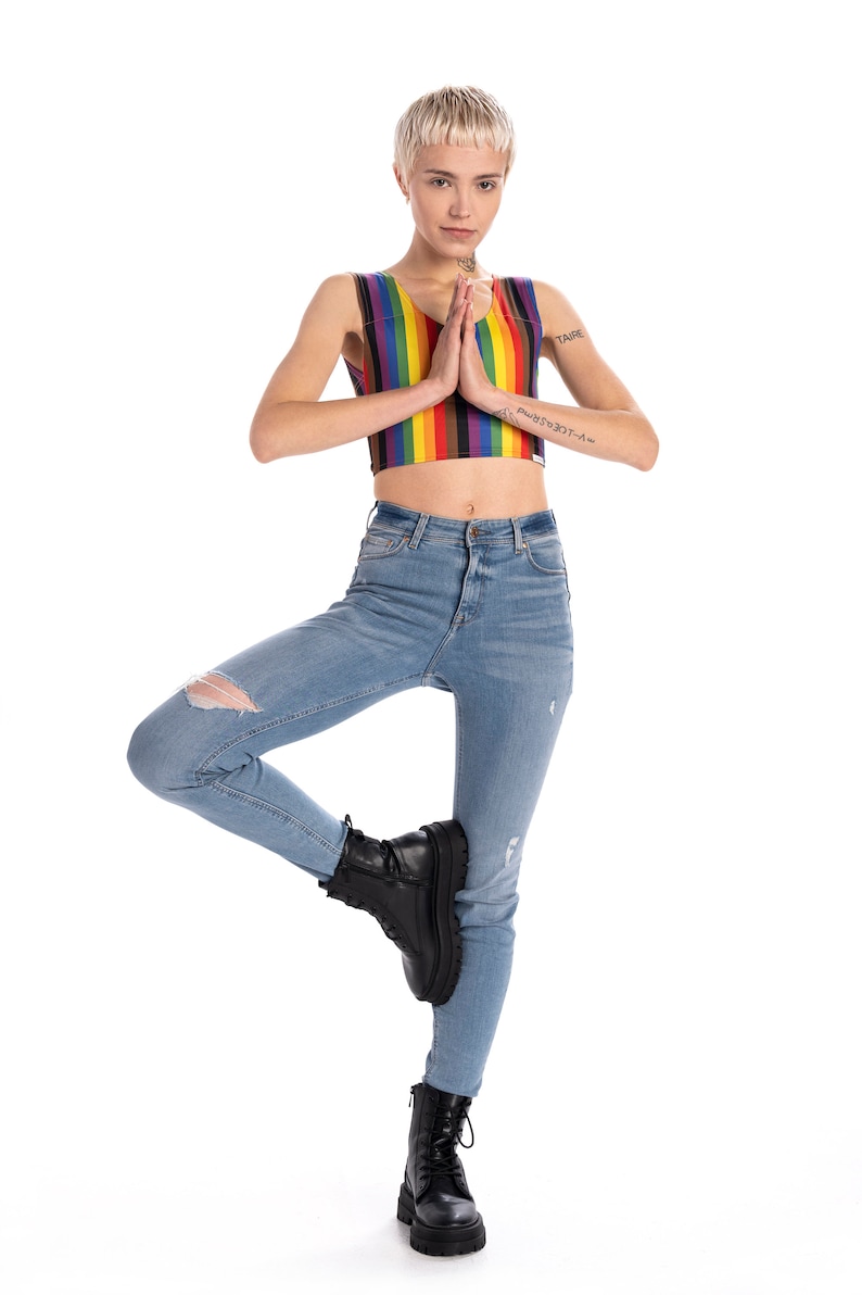 May include: A person wearing a rainbow striped crop top and blue jeans with rips. They are standing on one leg with their other leg bent at the knee and their foot resting on their thigh. They are wearing black combat boots and have tattoos on their arms. The text on their arm reads "TAIPEI" and "PLUS337-A".