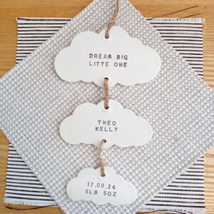 May include: White ceramic cloud-shaped ornaments with the text "Dream Big Little One", "Theo Kelly", and "17.09.24 6LB 5OZ" hanging from a string.