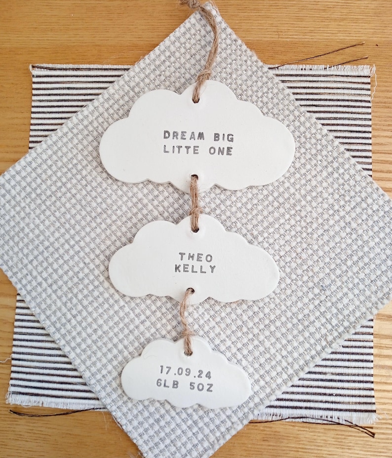 May include: White ceramic cloud-shaped ornaments with the text "Dream Big Little One", "Theo Kelly", and "17.09.24 6LB 5OZ" hanging from a string.