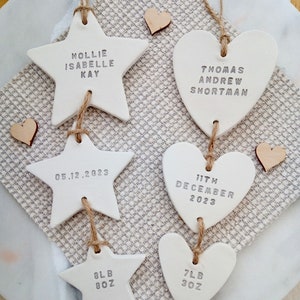 May include: White ceramic star and heart ornaments with personalized names and birth details. The star reads "Hollie Isabelle Kay" and "05.12.2023". The heart reads "Thomas Andrew Shortman" and "11th December 2023". Two additional ornaments read "8lb 8oz" and "7lb 3oz".