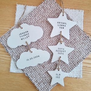 May include: Four white ceramic ornaments, two shaped like clouds and two shaped like stars, with black text. The cloud ornaments read "Jenson Thomas Willey" and "12.09.2024". The star ornaments read "Abigail Lorna Tew", "29th August 2024", and "9lb 10z".