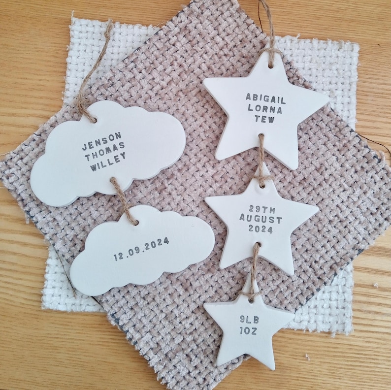 May include: Four white ceramic ornaments, two shaped like clouds and two shaped like stars, with black text. The cloud ornaments read "Jenson Thomas Willey" and "12.09.2024". The star ornaments read "Abigail Lorna Tew", "29th August 2024", and "9lb 10z".