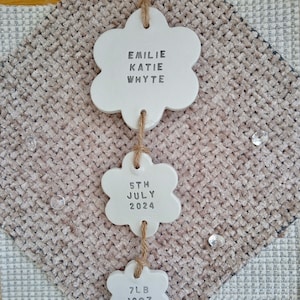 May include: White ceramic flower-shaped ornaments with the text "Emilie Katie Whyte", "5th July 2024", and "7lb 10oz" hanging from a string.