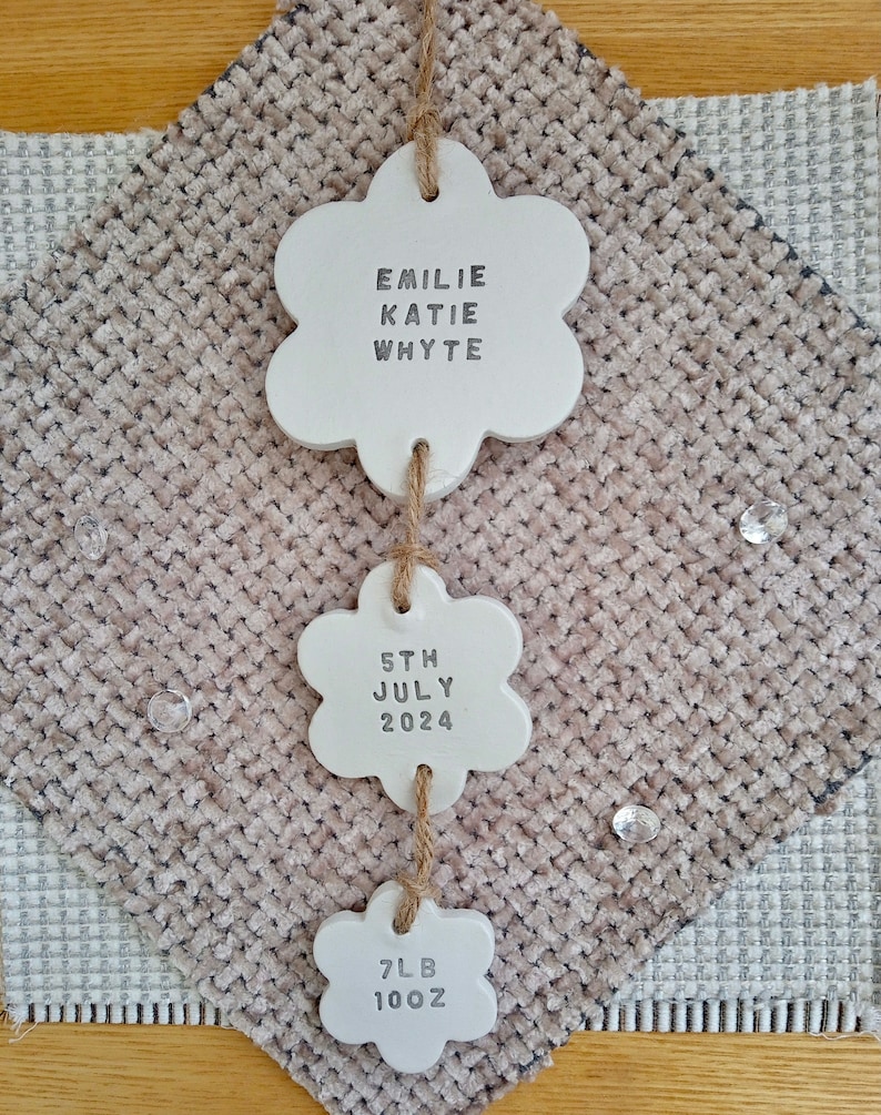 May include: White ceramic flower-shaped ornaments with the text "Emilie Katie Whyte", "5th July 2024", and "7lb 10oz" hanging from a string.