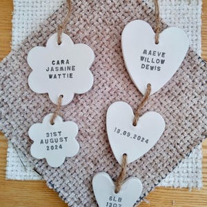 May include: Five white ceramic ornaments, two shaped like flowers and three shaped like hearts, hang from twine. The ornaments are decorated with black text. The flower ornaments read "Cara Jasmine Wattie" and "31st August 2024". The heart ornaments read "Maeve Willow Dewis", "18.09.2024", and "6lb 13oz".