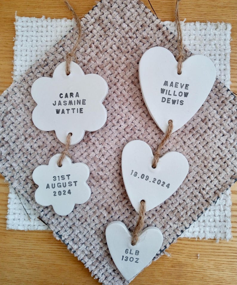 May include: Five white ceramic ornaments, two shaped like flowers and three shaped like hearts, hang from twine. The ornaments are decorated with black text. The flower ornaments read "Cara Jasmine Wattie" and "31st August 2024". The heart ornaments read "Maeve Willow Dewis", "18.09.2024", and "6lb 13oz".