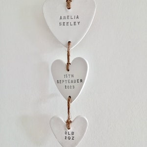 May include: Three white ceramic heart ornaments hanging from a twine string. The top heart says "Amelia Seeley", the middle heart says "15th September 2023", and the bottom heart says "8lb 2oz".