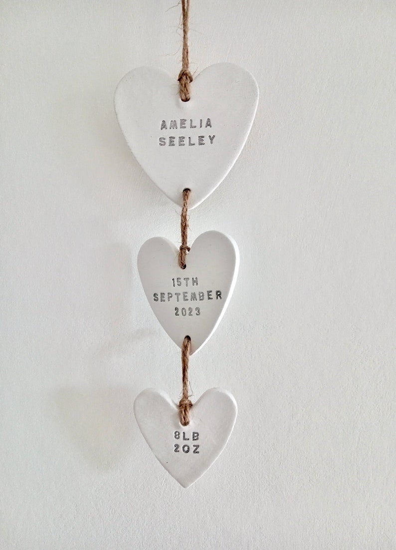 May include: Three white ceramic heart ornaments hanging from a twine string. The top heart says "Amelia Seeley", the middle heart says "15th September 2023", and the bottom heart says "8lb 2oz".