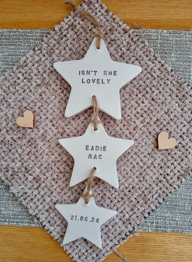 May include: A set of three white ceramic stars with twine hanging from each. The top star says "Isn't she lovely", the middle star says "Eadie Mac", and the bottom star says "21.06.24".