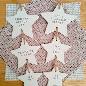 May include: Five white ceramic star ornaments with text. The top star reads "Arriety Monroe Day". The second star reads "Bodie Mcdonald Warner". The third star reads "09.07.2024 6.02AM". The fourth star reads "11th July 2024". The bottom left star reads "8LB 7OZ" and the bottom right star reads "9LB 10Z".