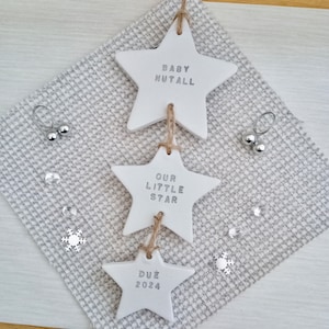 May include: Three white ceramic star ornaments with the text "Baby Nutall", "Our Little Star", and "Due 2024" hanging from a string.