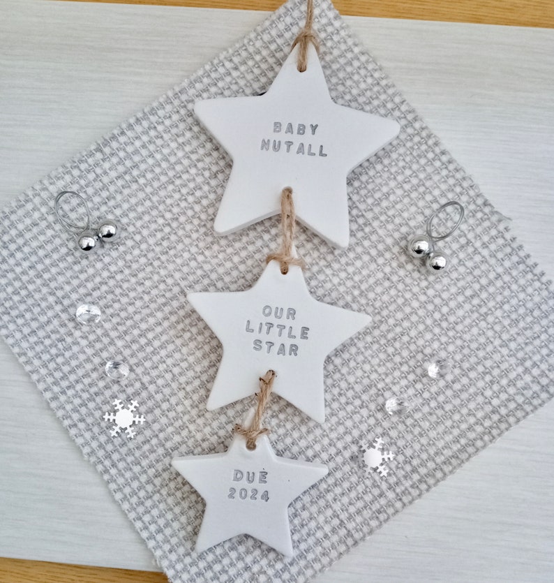May include: Three white ceramic star ornaments with the text "Baby Nutall", "Our Little Star", and "Due 2024" hanging from a string.