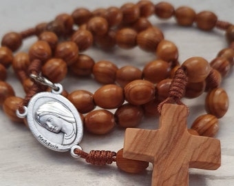 Può includere: Un rosario di legno con perline marroni e un ciondolo d'argento con l'immagine della Vergine Maria con il testo "Medjugorje". Il rosario include anche una croce di legno.