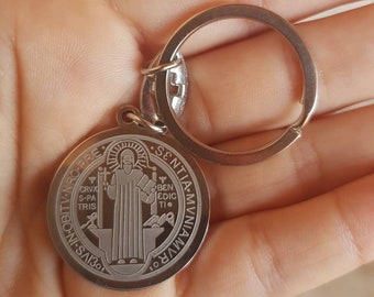 Può includere: Portachiavi in ​​argento con un ciondolo rotondo in metallo raffigurante San Benedetto. Il ciondolo ha le parole "S. BENEDICTUS" e "S. NTIA. MVN. VIA" attorno al bordo. Il ciondolo ha anche le parole "CRVX. S. PA. TRIS" e "BEN. EDIC. TI." all'interno del cerchio.