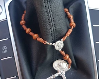 Könnte beinhalten: Ein braunes Holzrosenkranz mit einem silbernen Anhänger hängt am Schalthebel eines Autos. Der Rosenkranz ist auf einer braunen Schnur aufgefädelt.