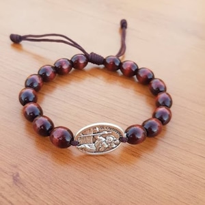 Puede incluir: Un brazalete de cuentas de madera marrón con un dije de plata que representa a San Cristóbal. El dije tiene la inscripción "St. Christopher".