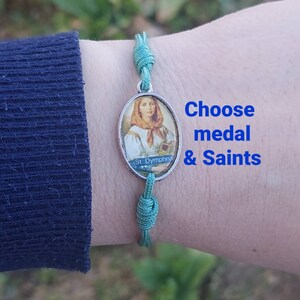 May include: A teal green cord bracelet with a silver oval charm featuring a painting of a saint. The charm has the text "St. Dymphna" on the bottom. The text "Choose medal & Saints" is written in blue on a white background.