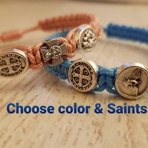 Könnte beinhalten: Zwei geflochtene Armbänder mit silbernen Anhängern. Ein Armband ist hellrosa und das andere hellblau. Die Anhänger umfassen ein Kreuz, einen Heiligen und eine Medaille. Der Text "Choose color & Saints" ist unterhalb der Armbänder sichtbar.