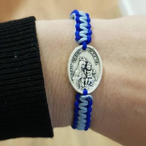 May include: A blue and gray braided bracelet with a silver charm featuring the Virgin Mary and baby Jesus. The charm has the text "ITALY VIRGIN OF CARMEL".