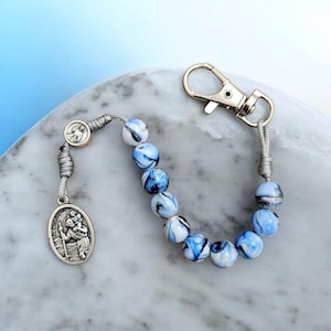 May include: A rosary-style keychain with blue and white marbled beads. It features a silver St. Christopher medal and a metal clasp. The beads are strung on a silver cord.