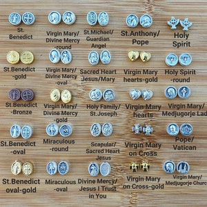 Puede incluir: Surtido de dijes de metal plateado y dorado con imágenes religiosas, incluyendo San Benito, la Virgen María, el Sagrado Corazón, la Sagrada Familia, y más. Los dijes se muestran sobre una superficie de madera.