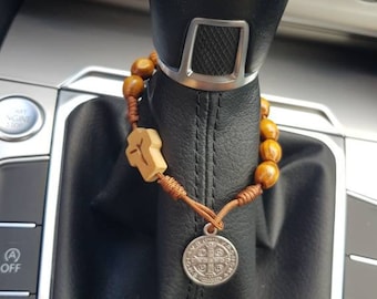 Könnte beinhalten: Ein braunes Holzrosenkranz mit einem silbernen Anhänger hängt am Schalthebel eines Autos. Der Rosenkranz hat ein Holzkreuz und einen silbernen Anhänger mit einem religiösen Design. Der Schalthebel ist aus schwarzem Leder und hat einen silbernen Schalthebelknauf mit den Zahlen 1, 2, 3, 4, 5, 6 und R.