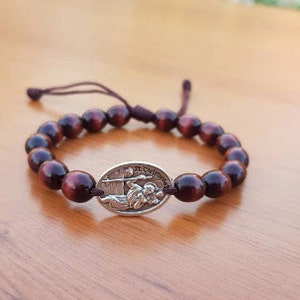 Puede incluir: Un brazalete de cuentas de madera marrón con un dije de plata que representa una figura religiosa. El dije tiene el texto "St. Christopher".