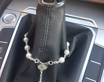 Könnte beinhalten: Ein silbernes Rosenkranz mit weißen Perlen hängt am Schalthebel eines Autos. Der Rosenkranz hat ein silbernes Kruzifix mit einer Jesus Christus-Figur.